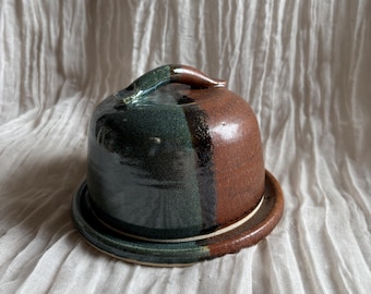 May include: A ceramic butter dish with a domed lid and a circular base. The lid is divided into two sections, one dark green and the other brown. A small handle is on top. The base is a matching color with a white rim.