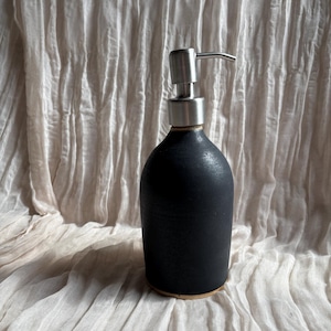 May include: A black ceramic soap dispenser with a stainless steel pump. The dispenser has a rounded shape and sits on a light brown base. The background is a textured, off-white fabric.