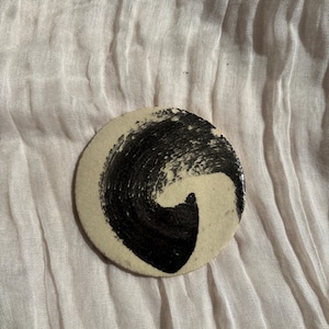 May include: A circular, cream-coloured coaster with a striking black abstract design. The design presents a swirling brushstroke pattern, creating a dynamic visual effect. The coaster is set against a soft, textured background.