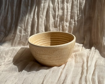 May include: A small, round, beige ceramic bowl with a textured, ridged interior. The bowl has a slightly flared rim and sits on a light-colored, textured fabric background. The bowl is likely handmade and suitable for serving food.