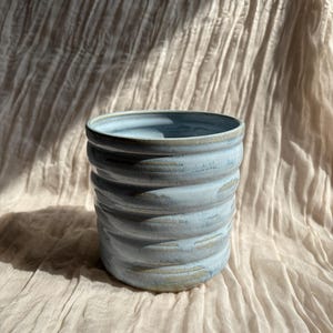 Könnte beinhalten: Eine handgefertigte Keramikvase mit hellblauer Glasur und einem strukturierten, geriffelten Design. Die Vase hat eine zylindrische Form und einen leicht ausgestellten Rand. Die Farbe ist ein weiches, gedämpftes Blau mit einem Hauch von Beige in den Rippen. Für die Inneneinrichtung.