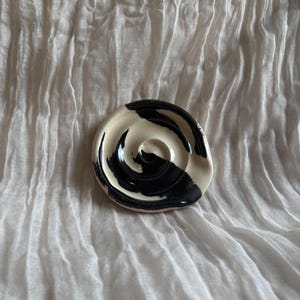 May include: A small, round ceramic dish with a cream-colored base and a bold, swirling black design. The dish has a shallow, concave shape and is likely handmade. The background is a textured, off-white fabric.
