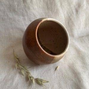 May include: A handcrafted ceramic bowl with a rounded form. The bowl displays a gradient of brown and tan tones, with a lighter rim. A sprig of dried grass is positioned beside the bowl, adding a natural element.