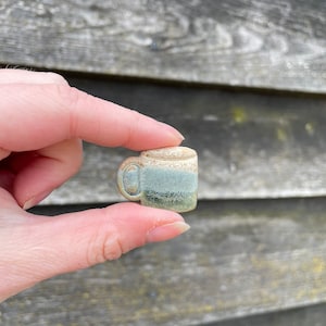 May include: A small ceramic mug with a light brown, green, and white glaze. The mug is hand-painted and has a unique, rustic look.