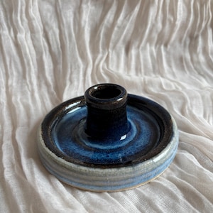 May include: A handcrafted ceramic candle holder with a dark blue interior and a circular base. The base features concentric rings of blue, grey, and brown. The candle holder is approximately 7.6 cm in diameter.