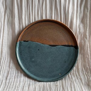 May include: A handcrafted ceramic plate with a two-tone design. The top half is a warm brown, and the bottom half is a deep teal. A thin black line separates the two colours. The plate is round and has a slightly raised edge.