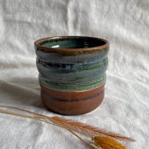 Könnte beinhalten: Eine handgefertigte Keramiktasse mit grüner und brauner Glasur. Die Tasse hat eine abgerundete Form und einen breiten Rand.