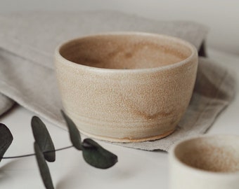 May include: Two ceramic bowls, one larger than the other, with a speckled beige glaze. The larger bowl is sitting on a linen napkin with a sprig of eucalyptus leaves.
