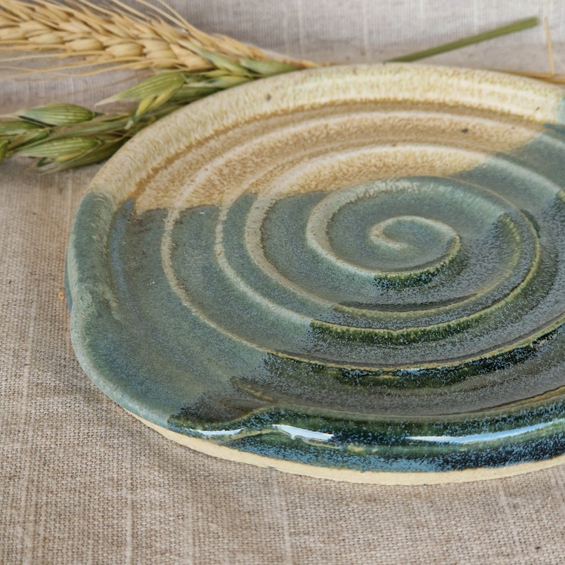 May include: A handmade ceramic spoon rest with a spiral design. The spoon rest features a blend of blue, green, and tan colours. The ceramic piece is round with a slightly raised edge and a textured surface.
