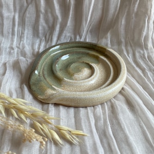 May include: A ceramic soap dish with a spiral design. The dish is a light beige colour with a green glaze in the centre. The dish is on a white, textured surface. Dried wheat stalks are in the lower left corner.