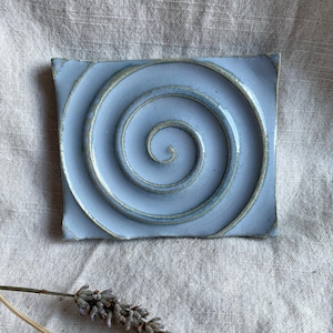 May include: A light blue ceramic tile with a raised spiral design. The tile has a slightly textured surface and a subtle green glaze.
