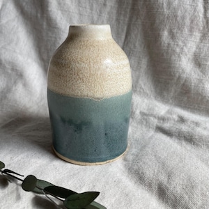 May include: A small ceramic vase with a white top and a teal blue bottom. The vase has a speckled glaze and a rounded shape.