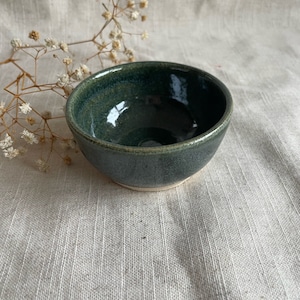 May include: A small, dark green ceramic bowl with a white rim. The bowl is sitting on a white linen fabric.