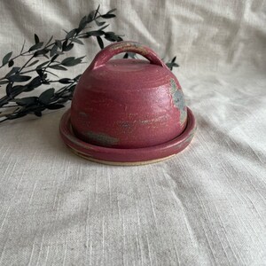 May include: A red ceramic butter dish with a lid and a handle. The dish has a rustic, handmade look with a speckled glaze.