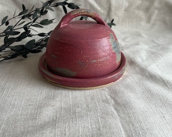 May include: A red ceramic butter dish with a lid and a handle. The dish has a rustic, handmade look with a speckled glaze.