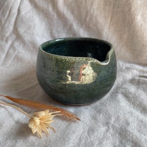 Peut inclure: Un petit bol en céramique vert foncé avec un bord légèrement irrégulier. Le bol a un glaçage moucheté et une subtile brillance métallique.