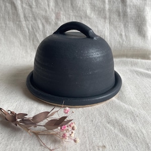 Könnte beinhalten: Eine schwarze Keramik-Butterdose mit Deckel und Griff. Die Dose steht auf einer weißen Stoffoberfläche.