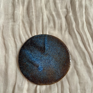 May include: A round, ceramic coaster with a speckled blue and brown glaze. The coaster has a dark blue center that fades to brown around the edges. The coaster is set against a white, textured fabric background.