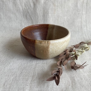 May include: A small, brown and white ceramic bowl with a speckled glaze. The bowl is sitting on a light brown fabric surface.