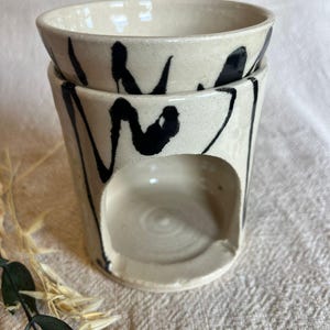 May include: A two-piece ceramic oil warmer with a cream-colored base and black abstract brushstrokes. The top bowl sits atop a cylindrical base with a circular opening. The warmer is designed for aromatherapy and home fragrance.