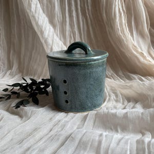 May include: A teal ceramic butter dish with a lid and handle. The cylindrical container has three small holes and sits on a white cloth. The background features a neutral-toned fabric.