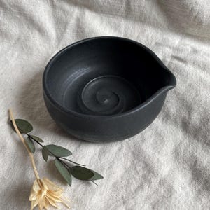 May include: A black ceramic bowl with a rounded lip and a spiral design on the inside. The bowl is sitting on a light beige fabric surface.