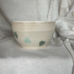 May include: A white ceramic bowl with a speckled glaze and green leaf designs. The bowl is round and has a wide rim.