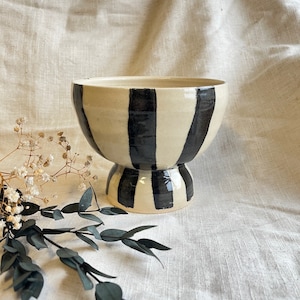 May include: A white ceramic bowl with black vertical stripes. The bowl has a pedestal base and is ideal for serving or displaying decorative items.