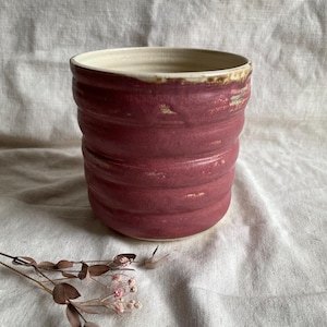 May include: A deep red ceramic planter with a white rim and a textured, wavy design. The planter is sitting on a white fabric surface.