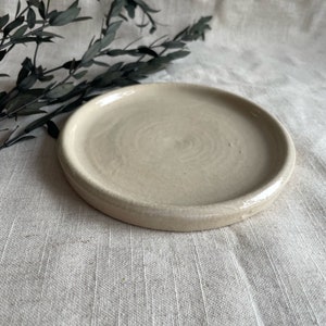 Puede incluir: Un peque&ntilde;o platillo de cer&aacute;mica de color blanco roto con un borde ligeramente elevado. El platillo tiene un sutil patr&oacute;n de remolino en la superficie.