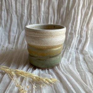 May include: A handcrafted ceramic mug with a distinctive, textured design. The mug has a layered colour scheme, shifting from a creamy white top to a golden-yellow middle, and ending with a muted green base. Dried wheat stalks are placed near the mug.