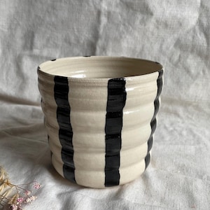 May include: A white ceramic planter with black vertical stripes. The planter has a rounded shape and a slightly flared rim.