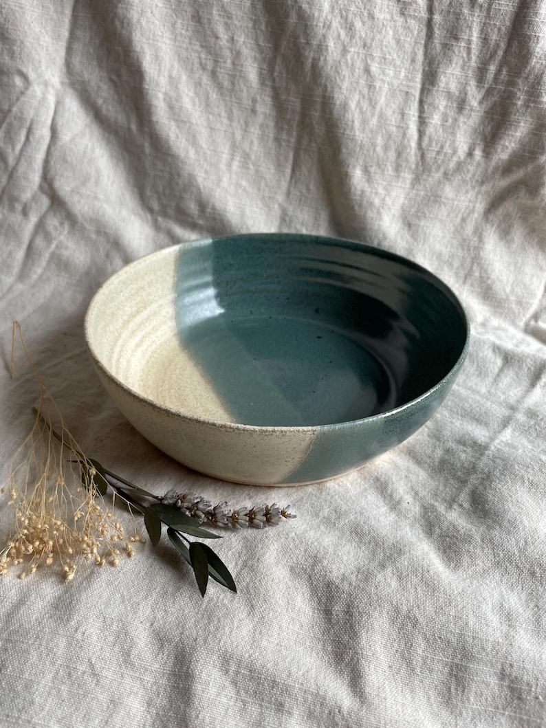 May include: A large, white and blue ceramic bowl with a speckled glaze. The bowl is sitting on a white fabric surface.