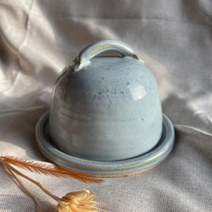 May include: A light blue ceramic butter dish with a lid and a handle. The dish has a speckled glaze and a white rim.