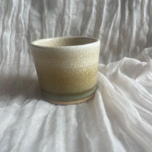 May include: A handcrafted ceramic cup with a speckled, textured finish. The cup displays a colour gradient, shifting from light beige at the top to a muted green at the base. The cup is set against a white, textured backdrop.