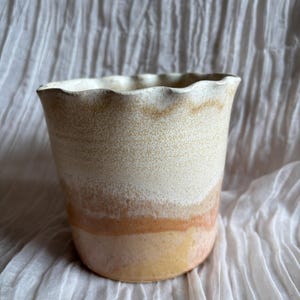 May include: A ceramic vase with a wavy rim. The vase has a speckled, off-white upper section, transitioning to a band of orange and then a yellow base. The background is a white, textured fabric.