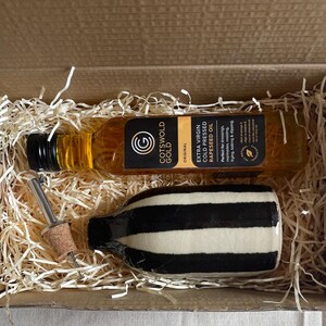 K&ouml;nnte beinhalten: Geschenkbox mit einer Flasche Cotswold Gold extra nativem kaltgepresstem Raps&ouml;l, einer schwarz-wei&szlig; gestreiften seifenf&ouml;rmigen Flasche und einem Flaschenverschluss. Die Box ist mit Stroh gef&uuml;llt.