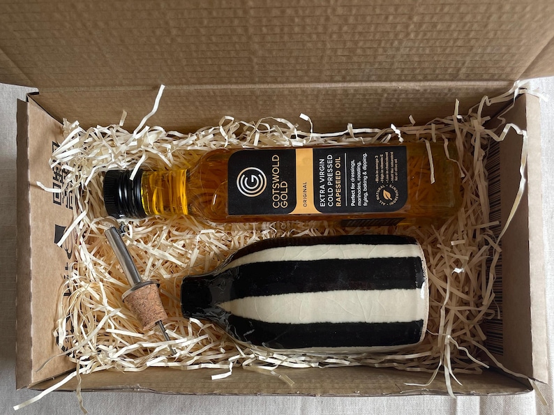 K&ouml;nnte beinhalten: Geschenkbox mit einer Flasche Cotswold Gold extra nativem kaltgepresstem Raps&ouml;l, einer schwarz-wei&szlig; gestreiften seifenf&ouml;rmigen Flasche und einem Flaschenverschluss. Die Box ist mit Stroh gef&uuml;llt.
