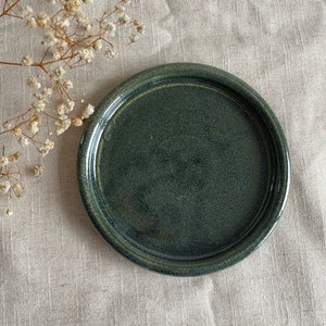 Könnte beinhalten: Ein dunkelgrüner Keramikteller mit leicht erhöhtem Rand. Der Teller hat eine gesprenkelte Oberfläche und ist perfekt zum Servieren kleiner Snacks oder Desserts.