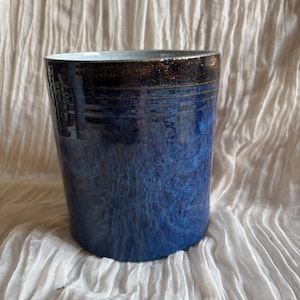 May include: A cylindrical ceramic vase in shades of blue. The top rim is dark brown with a textured surface. The vase has a speckled appearance and is likely handmade. The background is a white, textured fabric.