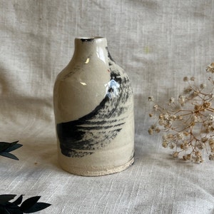 May include: A small, white ceramic vase with black brushstrokes. The vase has a rounded body and a narrow neck.