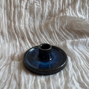 May include: A handcrafted ceramic candle holder. It features a dark blue interior and a black exterior. The candle holder has a circular base and a cylindrical centre for holding a candle. The glaze has a glossy finish.