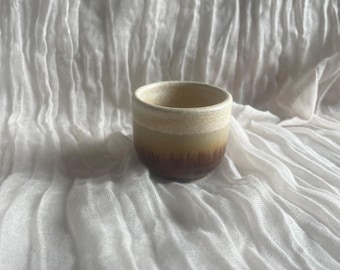 May include: A small, handcrafted ceramic cup with a gradient of colors. The top rim is a light beige, transitioning to a warm yellow and then a deep brown at the base. The cup has a simple, minimalist design.