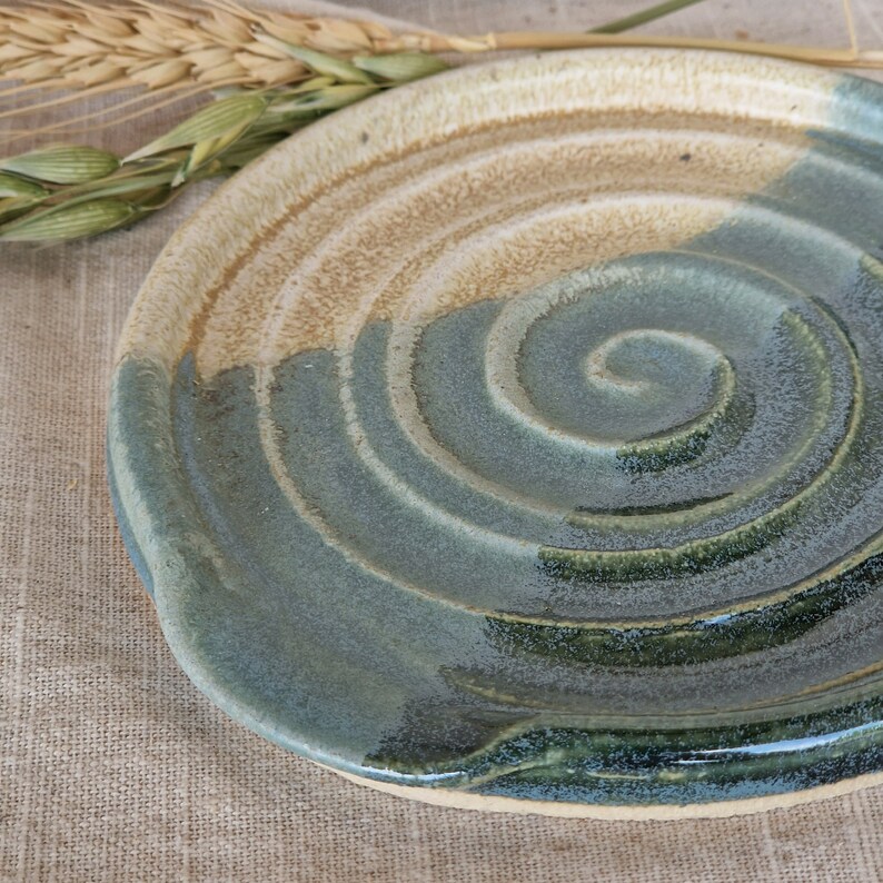 May include: A handmade ceramic plate with a spiral design. The plate features a two-tone colour scheme, with a beige upper section and a blue-green lower section. The plate's design is reminiscent of ocean waves.