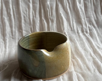 May include: A handmade ceramic bowl with a spout, featuring a blend of green and tan hues. The bowl has a rounded shape and a smooth, glazed finish. The pottery is set against a white, textured fabric background.