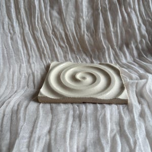 Könnte beinhalten: Ein quadratischer, cremefarbener Keramikuntersetzer mit einem Spiraldesign. Der Untersetzer hat ein erhabenes, konzentrisches kreisförmiges Muster. Der Hintergrund ist ein weißes, strukturiertes Gewebe.