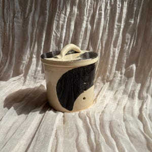 May include: A cream-coloured ceramic jar with a lid, adorned with a large black abstract design. The lid has a handle and a black rim. The jar is set against a textured, off-white fabric background.