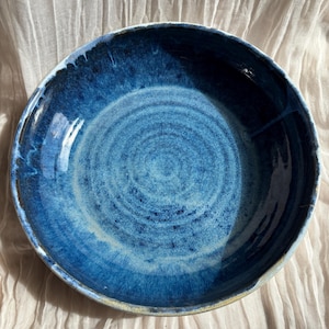 May include: A handmade ceramic bowl with a deep blue glaze. The bowl has a wide, shallow shape with a slightly uneven rim. The interior features concentric circles of lighter blue, creating a textured appearance. The bowl is suitable for serving or display.