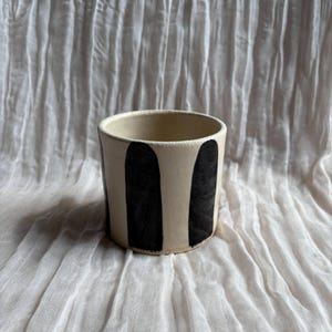 May include: A ceramic cup with a cream-colored base and vertical black painted stripes. The cup has a cylindrical shape and a smooth, unglazed exterior. The interior is a lighter cream color.