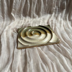 May include: Square ceramic coaster with a spiral design in shades of green, beige, and brown. The coaster has a textured surface and a slightly uneven edge. The coaster is placed on a light-coloured, textured fabric.